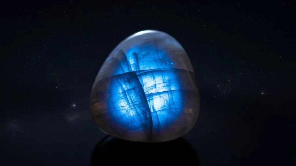 Una piedra luna pulida que muestra un intenso brillo azulado, conocido como adularescencia, sobre un fondo oscuro y estrellado, simbolizando misterio e intuición.