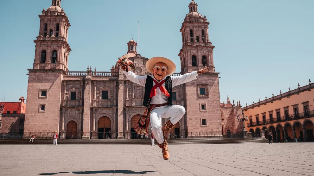 Bailarín ejecutando la Danza de los Viejitos con su máscara y traje típico en la plaza principal de Morelia, con la catedral de cantera rosa como fondo.